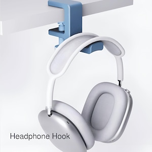 Puede incluir: Un gancho azul para auriculares con abrazadera fijado a una superficie blanca, que sostiene un par de auriculares blancos y grises. Los auriculares tienen una copa plateada y una diadema blanca. El texto "Headphone Hook" es visible.