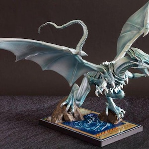 Könnte beinhalten: Detaillierte Figur eines hellblauen Drachen mit großen Flügeln, scharfen Krallen und einem langen, gebogenen Schwanz. Der Drache steht auf einer Basis mit Kartendesign, das komplizierte Details und eine dynamische Haltung zeigt. Sammlerstück.