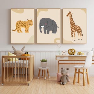 Op de afbeelding: Drie ingelijste dierenkunstdrukken. De prints tonen een luipaard, een olifant en een giraffe, allemaal in cartoonstijl. Het kunstwerk staat tegen een neutrale achtergrond met een beige rand. De kamer bevat ook een wieg, een bureau en knuffels.