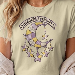 May include: A light yellow t-shirt featuring a graphic of a crescent moon and stars made of purple, yellow, and white checkered and floral quilt patterns. The text "STITCHING AFTER DARK" is printed in a dark purple arched font above the moon.