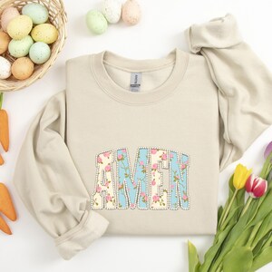 May include: A beige sweatshirt with the word "AMEN" in a floral design. The letters are outlined in white and set against a light blue background. Easter eggs and carrots are in the background.