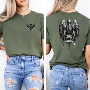 May include: Olive green t-shirt with a medical caduceus graphic on the front and a detailed angel medic design on the back. The angel medic design features wings, a helmet, and medical symbols. The shirt is paired with blue jeans.