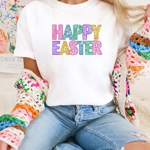 May include: White t-shirt with the text "HAPPY EASTER" in colorful, glittery letters. The shirt is paired with blue jeans and a colorful, crocheted cardigan. A casual and festive outfit.