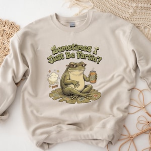 May include: Beige sweatshirt with a humorous graphic of a frog wearing sunglasses, holding a can of "Fizzy Farts", and sitting on a lily pad. The text reads "Sometimes I Just Be Fartin'!" A cartoon fart cloud is also present.