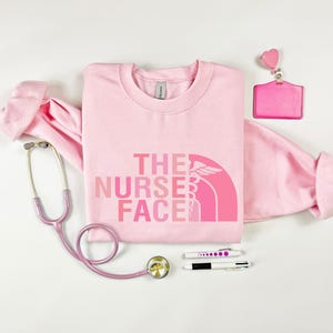a pink t-shirt with the text "THE NURSE FACE" printed on it, along with a stethoscope, a badge, and some pens.