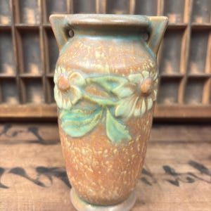 May include: A small, ceramic vase with a textured, speckled surface in shades of brown and tan. The vase features a floral design with green leaves and white flowers. It has two small handles and a rounded base.