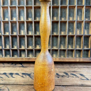 May include: A vintage wooden muddler, approximately 20 cm tall, with a rounded top and a bulbous base. The muddler is made of light-coloured wood and has a smooth, worn finish. It is set against a wooden background.