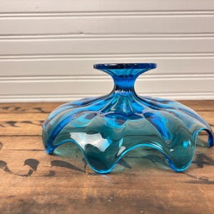 May include: A striking blue glass pedestal bowl with a ruffled edge. The bowl features a wide, wavy rim and a short, flared stem. The glass has a glossy finish, reflecting light. This decorative bowl is ideal for serving fruit or displaying decorative items.