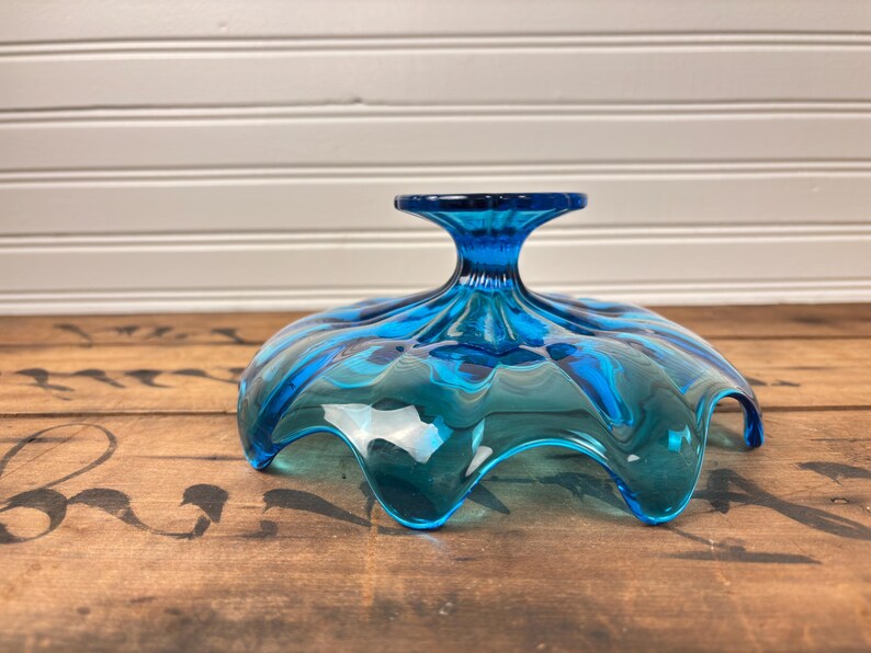May include: A striking blue glass pedestal bowl with a ruffled edge. The bowl features a wide, wavy rim and a short, flared stem. The glass has a glossy finish, reflecting light. This decorative bowl is ideal for serving fruit or displaying decorative items.
