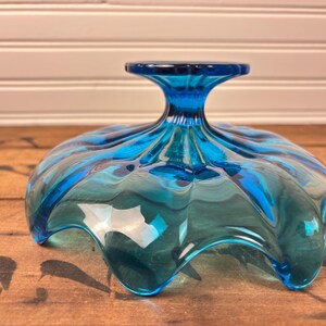 May include: A turquoise glass bowl with a pedestal base. The bowl has a ruffled, scalloped edge and a glossy finish. The bowl is on a wooden surface. The bowl's design is reminiscent of a flower.