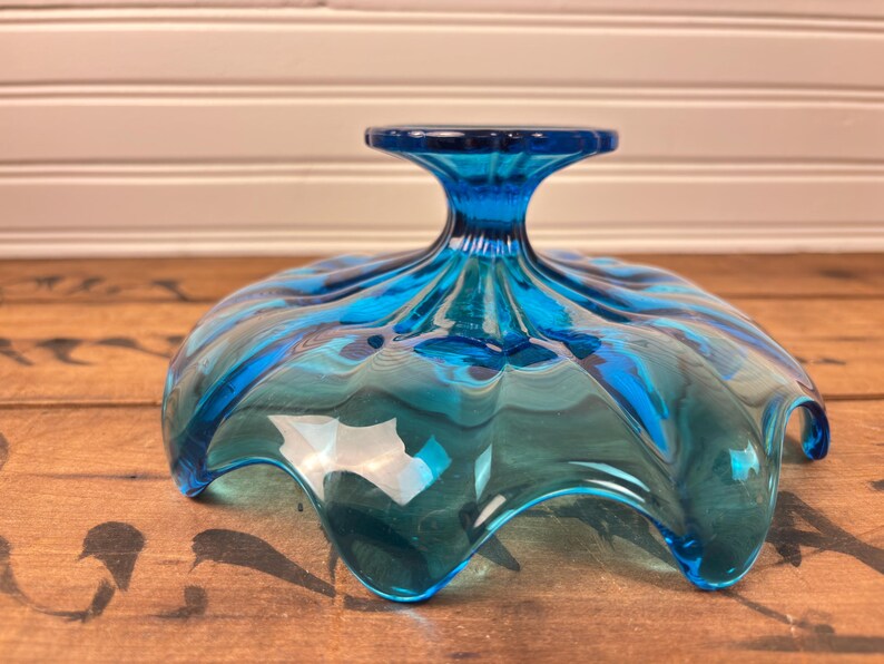 May include: A turquoise glass bowl with a pedestal base. The bowl has a ruffled, scalloped edge and a glossy finish. The bowl is on a wooden surface. The bowl's design is reminiscent of a flower.