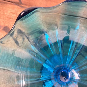 May include: A clear glass bowl in shades of blue and teal. The bowl features a distinctive, undulating rim and a central design that spreads outwards. The glass has a glossy finish, reflecting light. The bowl's design is eye-catching.
