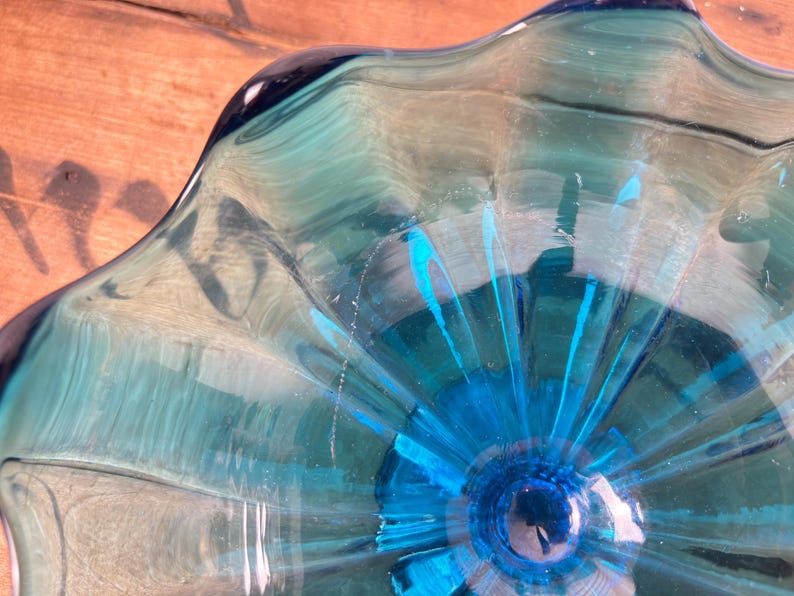 May include: A clear glass bowl in shades of blue and teal. The bowl features a distinctive, undulating rim and a central design that spreads outwards. The glass has a glossy finish, reflecting light. The bowl's design is eye-catching.