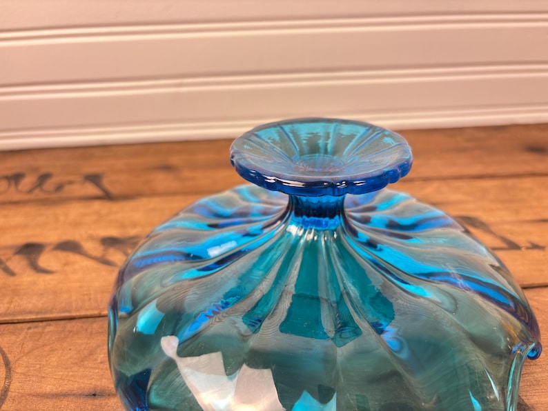 May include: A clear, turquoise glass bowl with a pedestal base. The bowl features a fluted design with a swirl pattern. The base is a solid, round shape. The bowl is likely for serving or display.