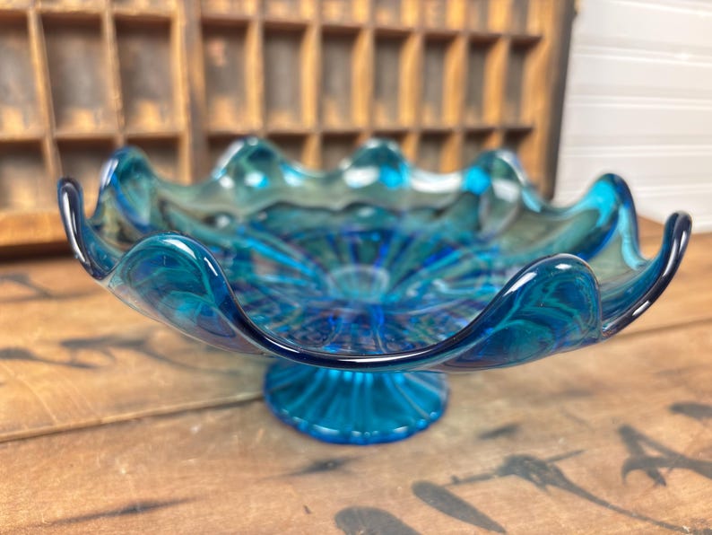 May include: A striking blue glass bowl with a scalloped edge and a pedestal base. The bowl features a swirling, textured design in shades of teal and blue. This decorative piece is suitable for serving or display.