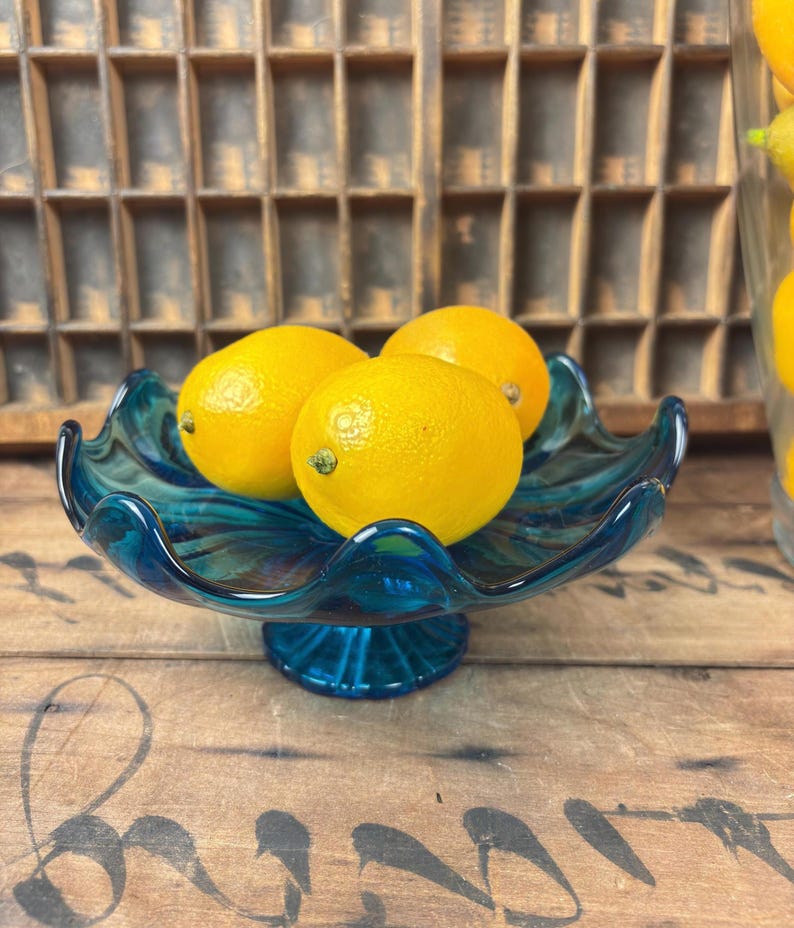 May include: A teal glass serving dish with a ruffled edge, holding three bright yellow lemons. The dish has a pedestal base and sits on a wooden surface. The lemons are round and have a textured appearance.