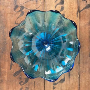 May include: A clear, teal-coloured glass bowl with a ruffled edge. The bowl has a deep blue centre with radiating streaks of blue. The bowl is sitting on a wooden surface.