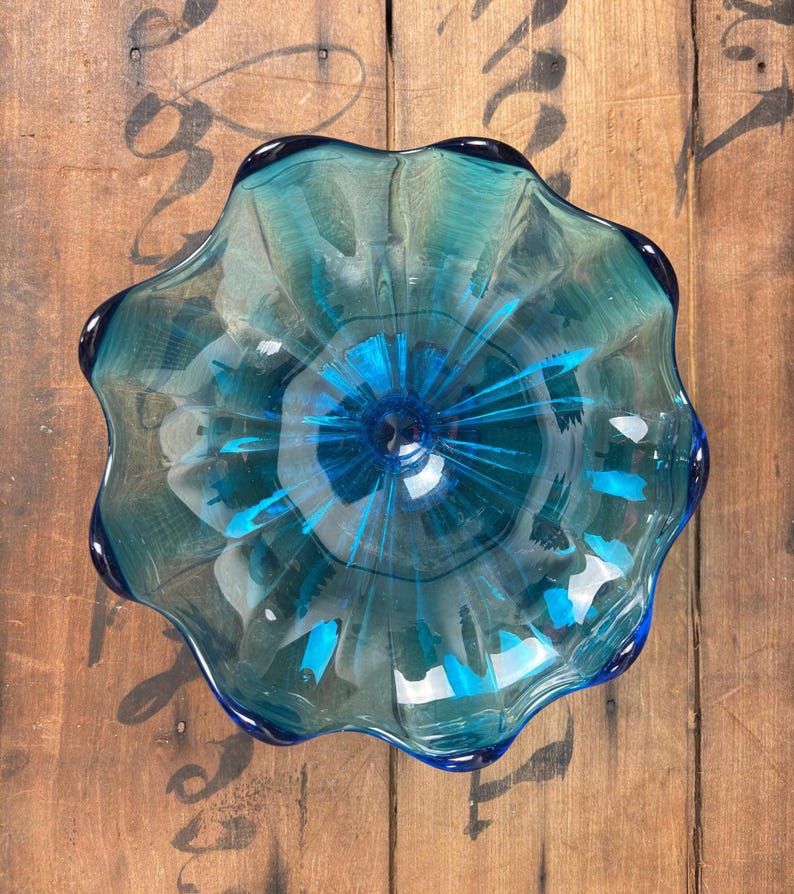 May include: A clear, teal-coloured glass bowl with a ruffled edge. The bowl has a deep blue centre with radiating streaks of blue. The bowl is sitting on a wooden surface.