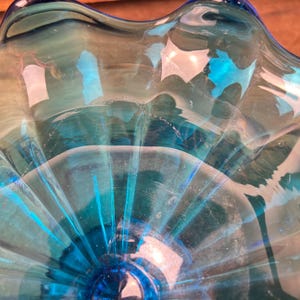 May include: A close-up of a clear, turquoise glass bowl. The bowl has a ruffled edge and a circular base. The glass has a rippled texture, and the light reflects off the surface, creating highlights and shadows.