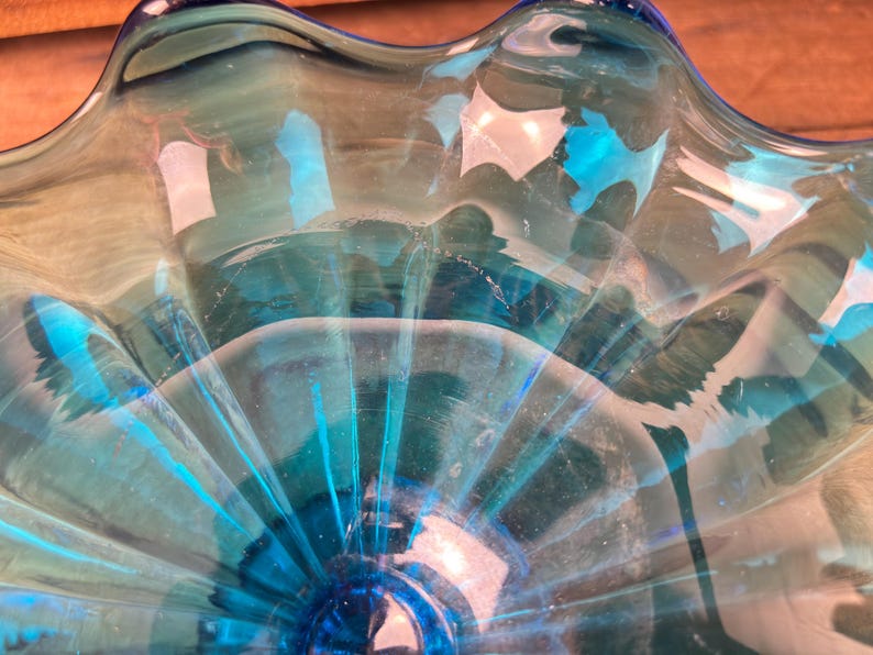 May include: A close-up of a clear, turquoise glass bowl. The bowl has a ruffled edge and a circular base. The glass has a rippled texture, and the light reflects off the surface, creating highlights and shadows.