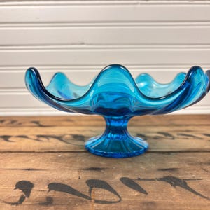 May include: A striking blue glass bowl with a wavy edge and a pedestal base. The bowl is made of translucent glass, displaying a swirling pattern of light and dark blue shades. This decorative bowl is ideal for displaying fruit or other items.