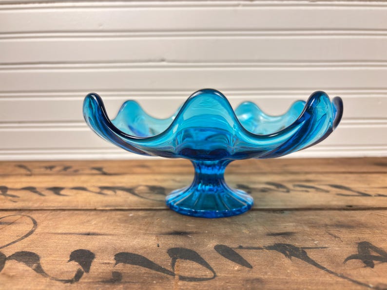 May include: A striking blue glass bowl with a wavy edge and a pedestal base. The bowl is made of translucent glass, displaying a swirling pattern of light and dark blue shades. This decorative bowl is ideal for displaying fruit or other items.