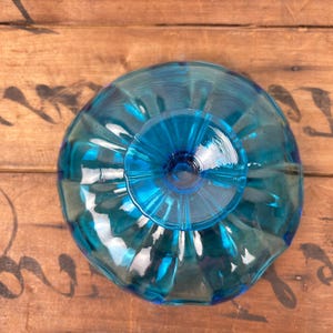 May include: A turquoise glass bowl with a fluted design. The bowl has a circular base and a wide, scalloped rim. The glass reflects light, creating a shimmering effect. The bowl is set against a wooden surface.