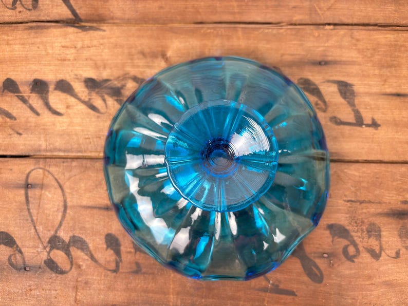 May include: A turquoise glass bowl with a fluted design. The bowl has a circular base and a wide, scalloped rim. The glass reflects light, creating a shimmering effect. The bowl is set against a wooden surface.