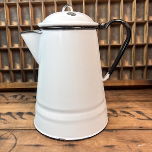 May include: White enamelware coffee pot with a black handle and rim. The pot has a lid with a small handle and a spout for pouring. The pot is sitting on a wooden surface.