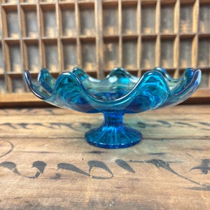 May include: A striking blue glass pedestal bowl with a wavy rim. The bowl features a circular base and a flared, scalloped edge. The glass has a marbled effect. The bowl is displayed on a wooden surface.