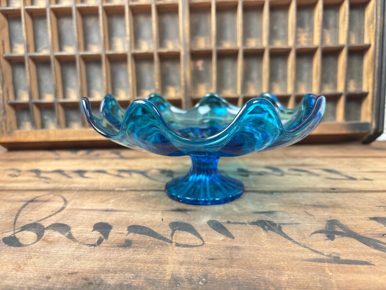 May include: A striking blue glass pedestal bowl with a wavy rim. The bowl features a circular base and a flared, scalloped edge. The glass has a marbled effect. The bowl is displayed on a wooden surface.