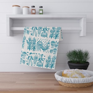 May include: A cream-coloured tea towel with a teal folk art pattern. The design includes figures, plants, and a rooster. The towel hangs on a white shelf above a wooden surface.