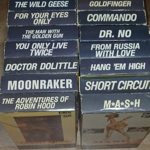 Peut inclure: Une pile de cassettes VHS vintage avec des titres tels que "Thunderball" et "Goldfinger". Les boîtiers sont principalement bleu foncé avec du texte blanc, et certains présentent des extraits de films. Les cassettes sont empilées.