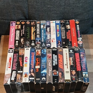 Peut inclure: Une collection de cassettes VHS avec des titres de films tels que "Jerry Maguire", "Independence Day" et "Speed". Les cassettes sont empilées sur une surface en bois, présentant une variété de couvertures de films colorées des années 1990 et du début des années 2000.