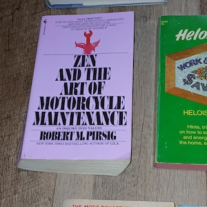 K&ouml;nnte beinhalten: Eine Sammlung von Vintage-B&uuml;chern mit farbenfrohen Einb&auml;nden. Das zentrale Buch, "Zen and the Art of Motorcycle Maintenance", hat einen hellvioletten Einband mit fettgedrucktem wei&szlig;em Text. Weitere B&uuml;cher sind "Feathers" und "Heloise".