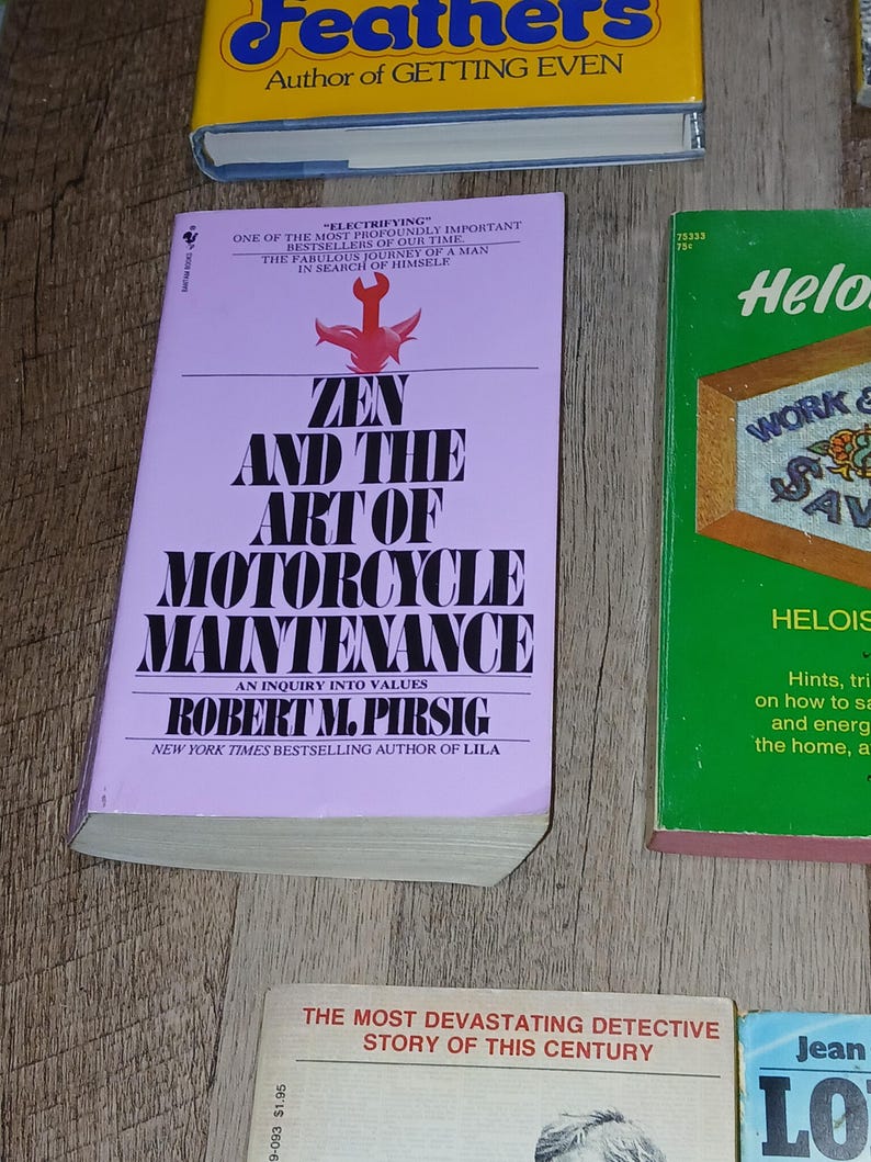 K&ouml;nnte beinhalten: Eine Sammlung von Vintage-B&uuml;chern mit farbenfrohen Einb&auml;nden. Das zentrale Buch, "Zen and the Art of Motorcycle Maintenance", hat einen hellvioletten Einband mit fettgedrucktem wei&szlig;em Text. Weitere B&uuml;cher sind "Feathers" und "Heloise".