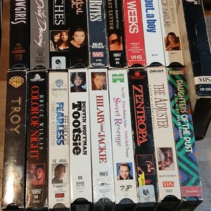 Può includere: Una collezione di videocassette VHS con titoli di film come "Showgirls", "Dirty Dancing" e "Beaches". Le cassette presentano locandine di film e sono di vari colori e design.