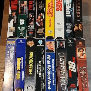 Puede incluir: Una colección de cintas VHS con títulos de películas como "Desperado", "Robin Hood", "Star Wars: La Amenaza Fantasma" y "Sneakers". Las cintas son de varios colores y presentan ilustraciones y títulos de películas.