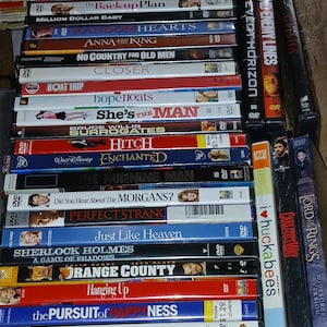 May include: A stack of DVDs with various movie titles visible, including "The Hangover," "Pride & Prejudice," and "Hitch." The cases are in various colors, with text and images of the movie stars. Some DVDs have price stickers.