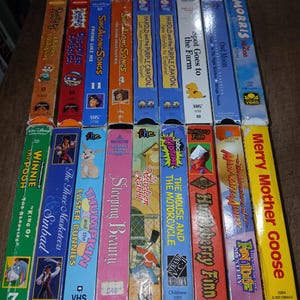 May include: A collection of vintage VHS tapes featuring animated movies and children's programming. Titles include "Winnie the Pooh," "The Three Musketeers," "Sleeping Beauty," and "The Mouse and the Motorcycle." The tapes are in various colors with colorful cover art.