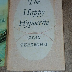 K&ouml;nnte beinhalten: Ein Vintage-Buchcover mit dem Titel "The Happy Hypocrite" in eleganter Schrift. Der Name des Autors, Max Beerbohm, ist ebenfalls zu sehen. Das Cover hat einen hellpfirsichfarbenen Hintergrund mit einem ovalen Design und roten Linien.