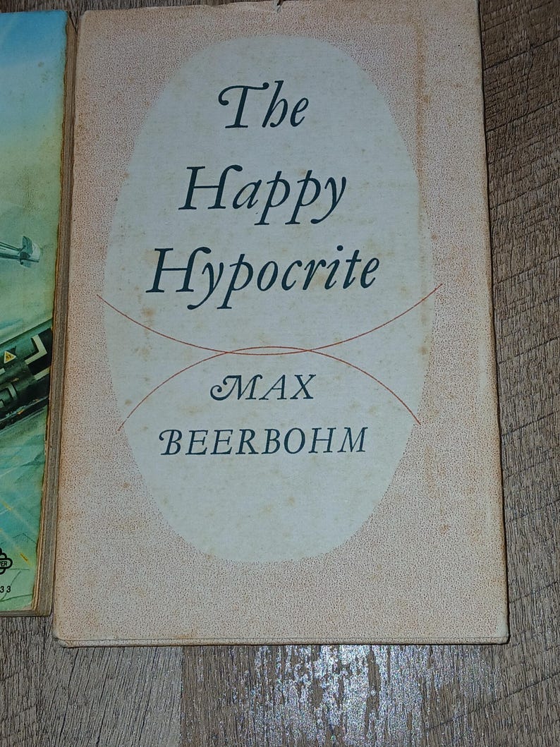K&ouml;nnte beinhalten: Ein Vintage-Buchcover mit dem Titel "The Happy Hypocrite" in eleganter Schrift. Der Name des Autors, Max Beerbohm, ist ebenfalls zu sehen. Das Cover hat einen hellpfirsichfarbenen Hintergrund mit einem ovalen Design und roten Linien.