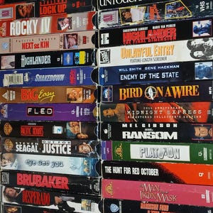 Peut inclure: Une pile de cassettes VHS vintage avec des titres de films tels que "Rocky IV", "Highlander" et "The Untouchables". Les cassettes présentent des étiquettes colorées avec des illustrations et du texte de films, représentant divers genres.