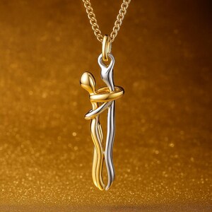 Puede incluir: Un collar con colgante de oro y plata que presenta un diseño abstracto de dos figuras abrazándose. El colgante cuelga de una cadena dorada. Las figuras están entrelazadas, una en oro y otra en plata, creando un contraste sobre un fondo dorado.