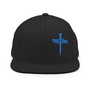 Puede incluir: Gorra de béisbol negra con visera plana. La gorra presenta un diseño de cruz azul y el texto "TOUGH AS NAILS". Fabricada con material resistente, tiene un ajuste estructurado.