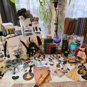 May include: A collection of gothic-themed items, including tote bags, journals, candles, and decorative objects. The items are arranged on a table with a floral tablecloth, with a variety of colors and textures.