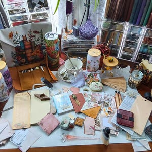 May include: A collection of stationery and craft supplies including a floral book-themed tote bag, several glass tumblers with bamboo lids, various notebooks, small envelopes, decorative paper scraps, a glass dip pen, and a small red leather wallet.