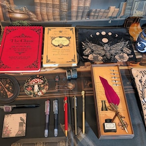 May include: A collection of stationery items including a red spiral notebook, a yellow book, a black pouch with a moth print, a quill pen set with ink, a book titled Tales Buried Deep, various pens, a small round frame, and decorative metal trinkets.