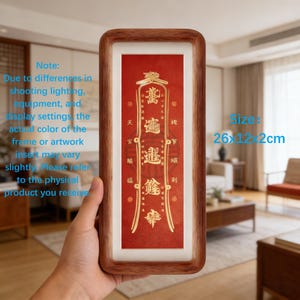 May include: A rectangular wooden framed artwork with a red background and gold text. The artwork measures 10.2 x 4.7 x 0.8 inches. The frame is a warm brown color. The artwork has Chinese characters and is held by a person.