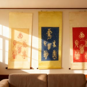 Puede incluir: Tres rollos de pared de estilo asiático con caligrafía e ilustraciones. Los rollos son en tonos de amarillo, azul y rojo. La caligrafía es en tinta negra y roja. Los rollos cuelgan de una pared blanca sobre un sofá beige.